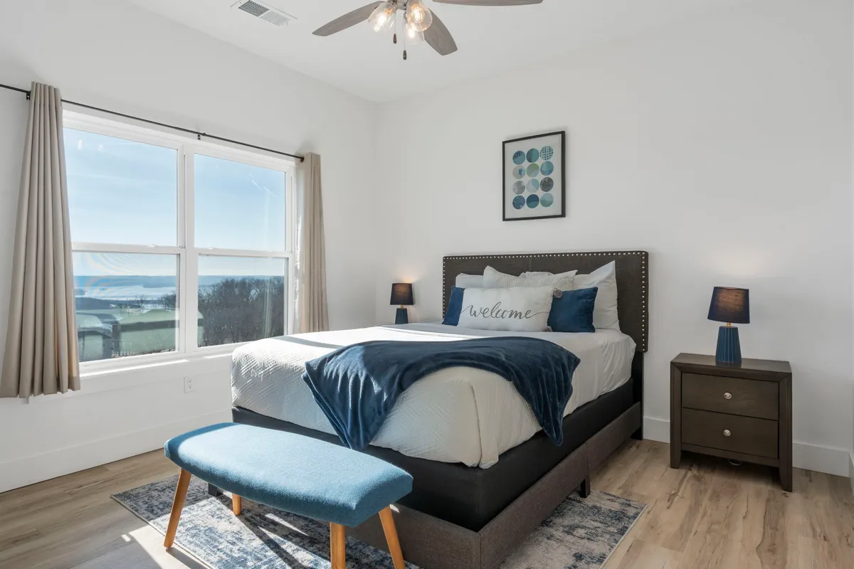 A serene bedroom with a bed adorned with decorative pillows, large windows that let in natural light, nightstands with lamps, and a bench at the foot of the bed. Soft neutral tones and inviting decor create a tranquil, restful atmosphere.