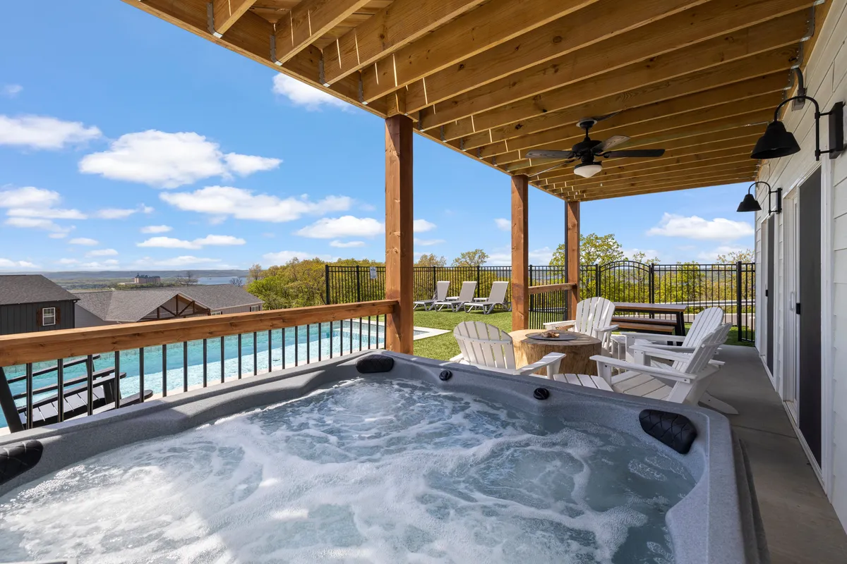 The image showcases the exterior of a spacious, upscale home in Branson, Missouri. In the foreground, a sparkling private pool and bubbling hot tub invite relaxation, while an outdoor seating area beneath a stylish wooden ceiling offers space for gathering. The scene is bathed in tranquil sunset light, evoking a peaceful, resort-like atmosphere ideal for group getaways.