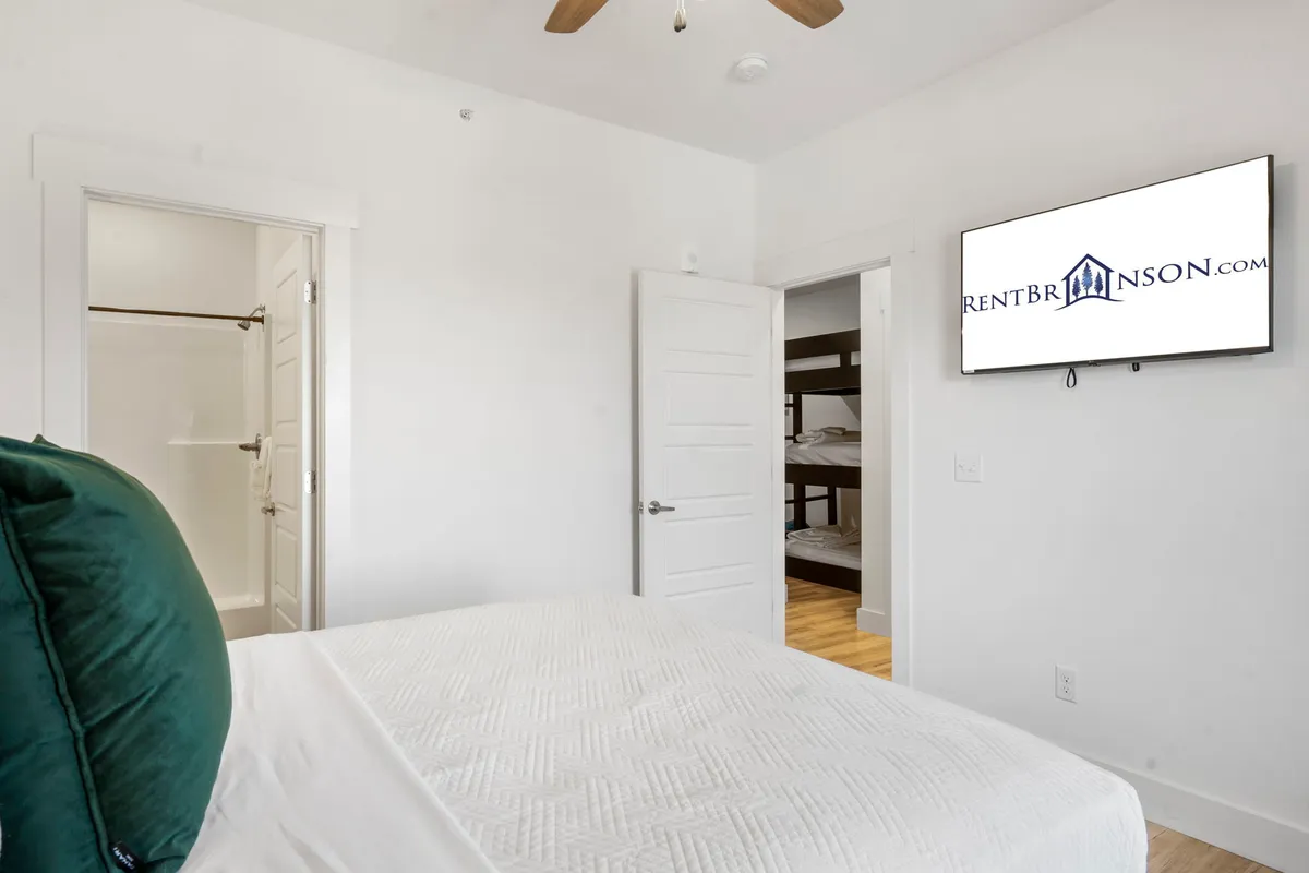 A spacious bedroom featuring a comfortable bed with crisp white bedding in the foreground, a set of bunk beds in the background, and a wall-mounted TV. The room feels modern and welcoming, providing options for both adults and children to relax or enjoy a movie night together.