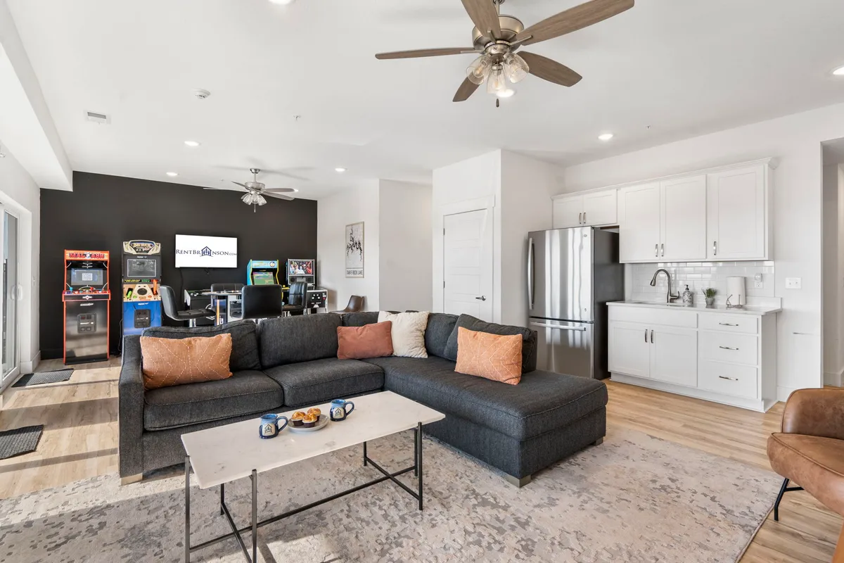 This inviting interior scene showcases a large sectional sofa positioned in front of a set of arcade machines, with a kitchenette and coffee table topped with snacks visible in the background. The mood is casual and lively, clearly designed to bring people together for game nights, movie watching, or intimate gatherings.