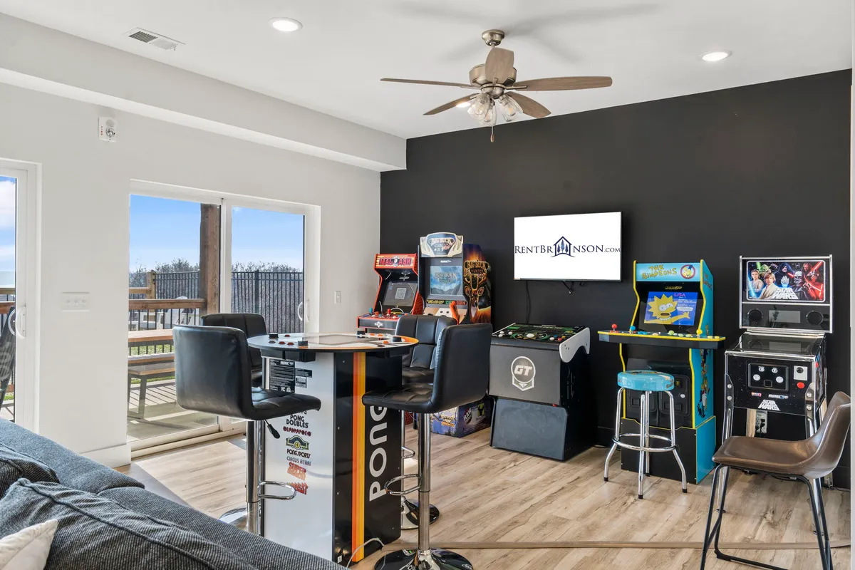 A vibrant interior space filled with several arcade machines lines the wall. A bar table and ceiling fan add to the casual, game room ambiance, inviting guests of all ages to engage in friendly competition or simply hang out and relax.