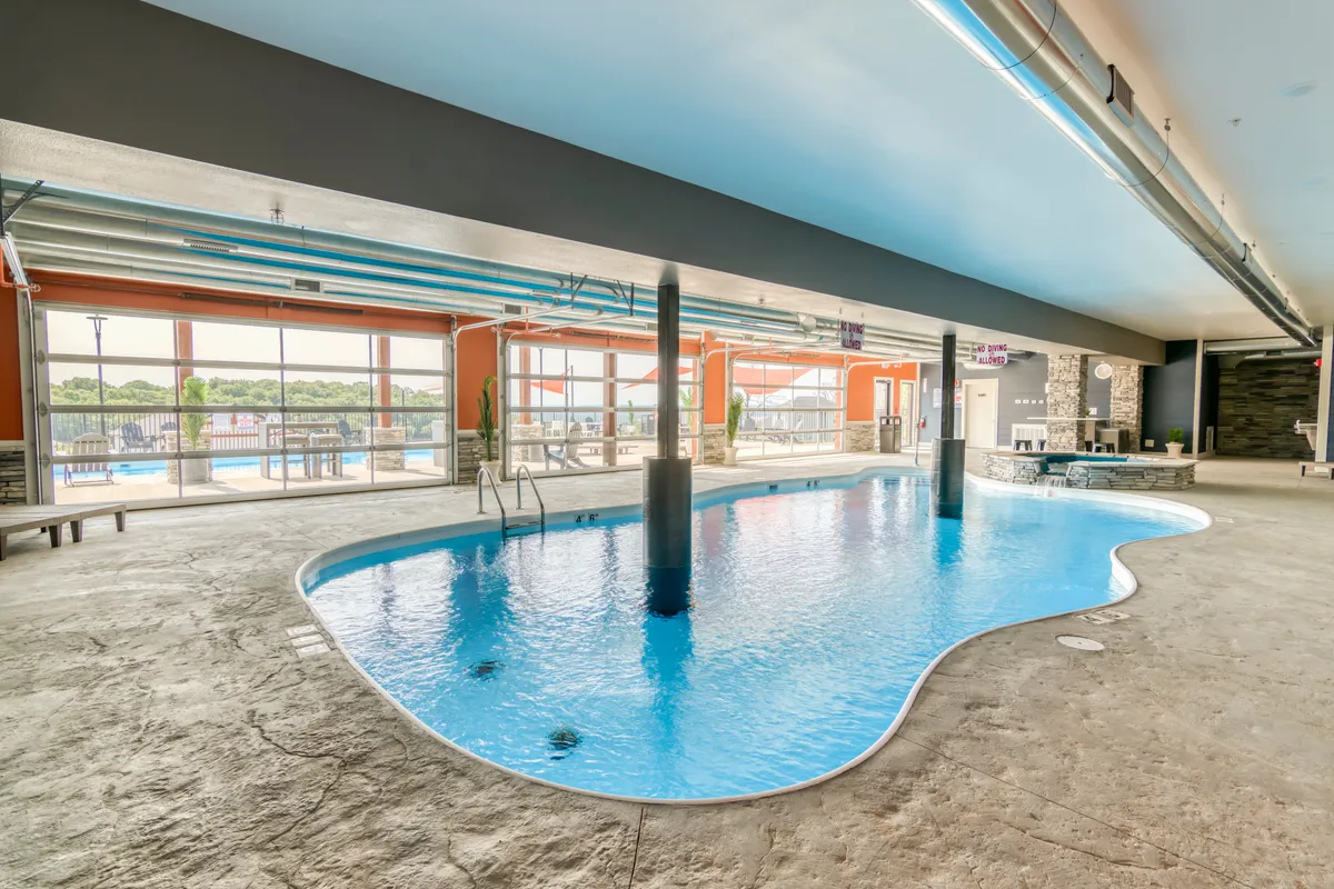 An interior view of a community indoor pool, featuring large windows that let in natural light and a comfortable seating area nearby. The tranquil blue water and clean, spacious environment invite guests to swim or relax regardless of the season.