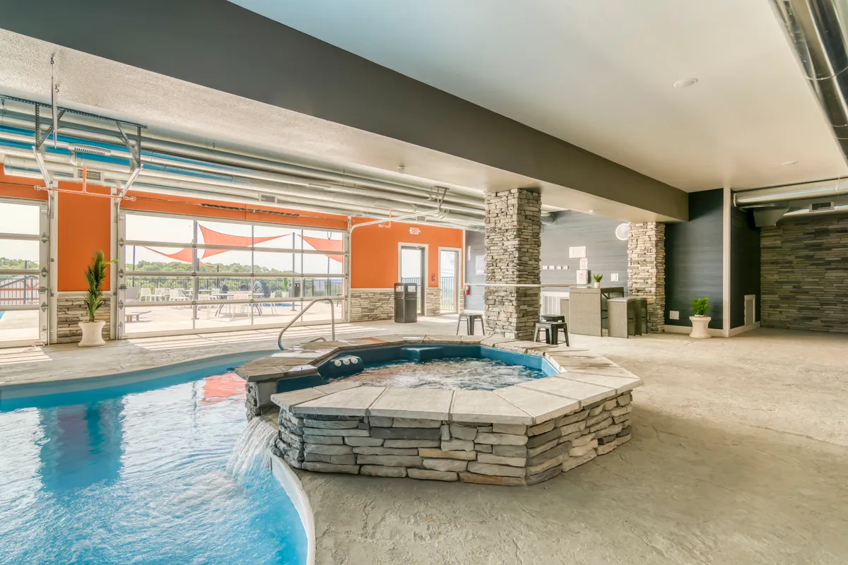 A tranquil indoor pool area features a hot tub, large windows letting in natural light, and stone features that give the space a cozy, inviting feel. The pool is ideal for swimming or relaxing regardless of the weather outside.