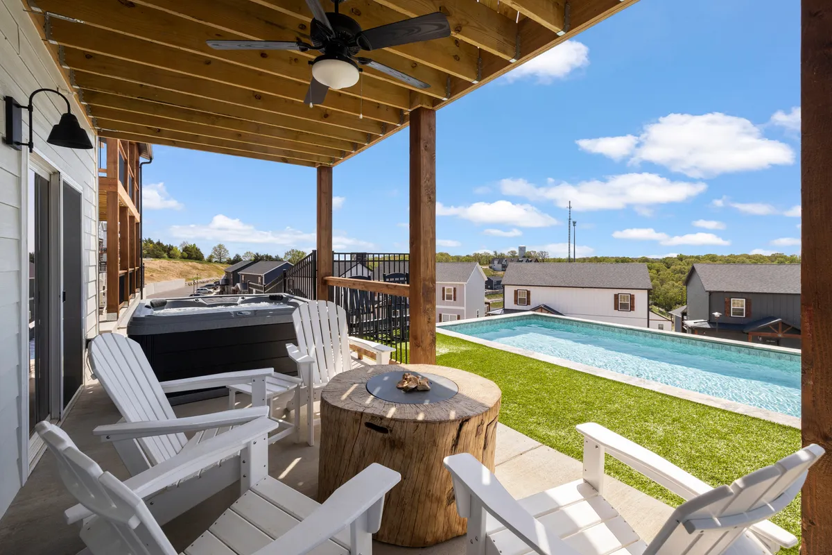 An exterior view featuring a sparkling swimming pool and an adjacent bubbling hot tub. Wooden Adirondack chairs, a table, and a relaxed patio set-up create a welcoming atmosphere for both social gatherings and peaceful moments by the water. The firepit is subtly visible, hinting at cozy evening gatherings.