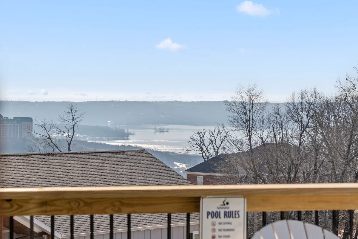 A peaceful overview of a pool area with a scenic water view and bare trees in the background, flanked by rooflines of neighboring houses. The pool rules sign and arrangement suggest a relaxing, social-friendly environment, with natural beauty all around.